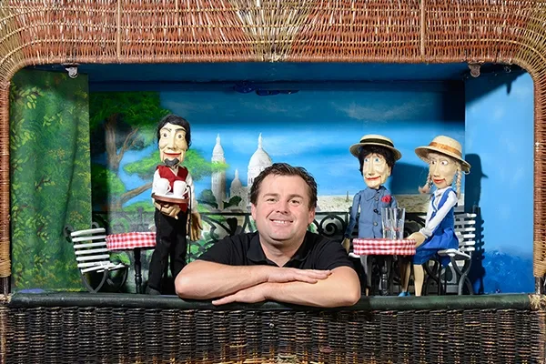 Alfred Büttner lächelnd an der Weidenbühne des Korbtheaters, im Hintergrund seine Puppen in einer Café-Szene